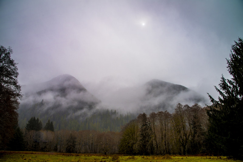 Two mountain peaks emerging from fog with a diffused sun above an overcast meadow foreground